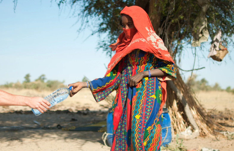 A woman in a colorful, patterned dress and red headscarf accepts a plastic water bottle from an outstretched hand, standing near a tree in a dry, rural area