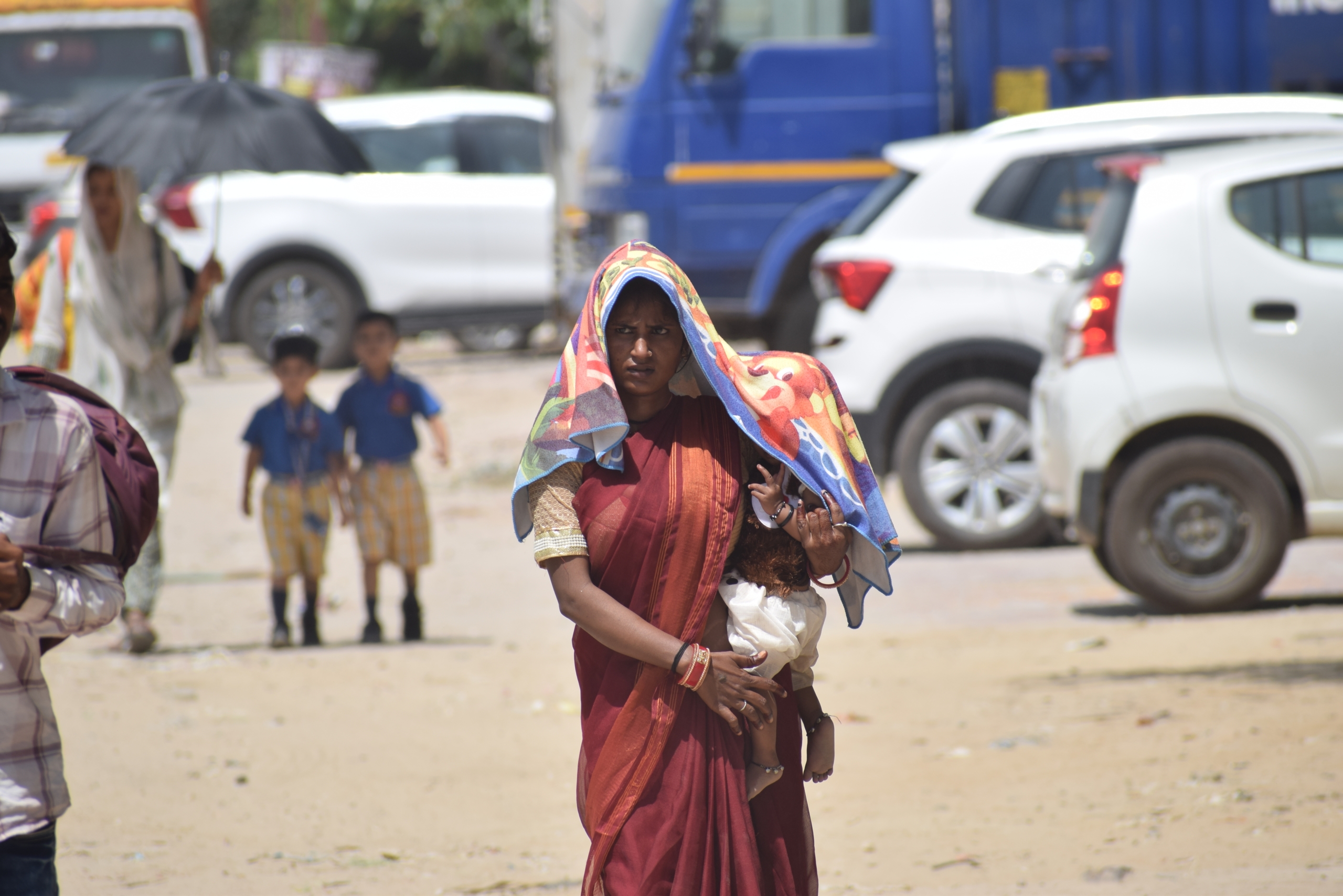Woman walking in the heat with a child, using a cloth to shield herself from the sun, on a busy street with cars and pedestrians."
