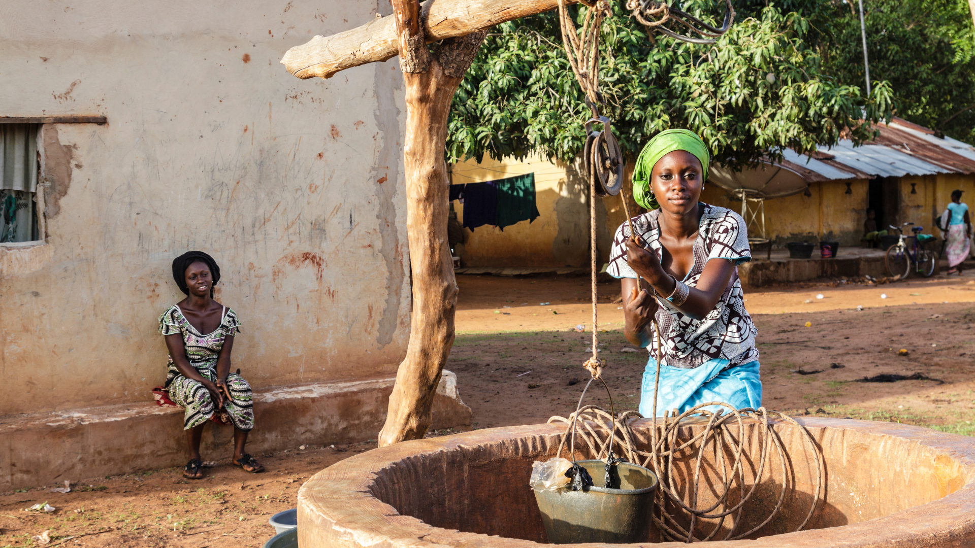 Woman retrieving water from a well while another woman watches smiling