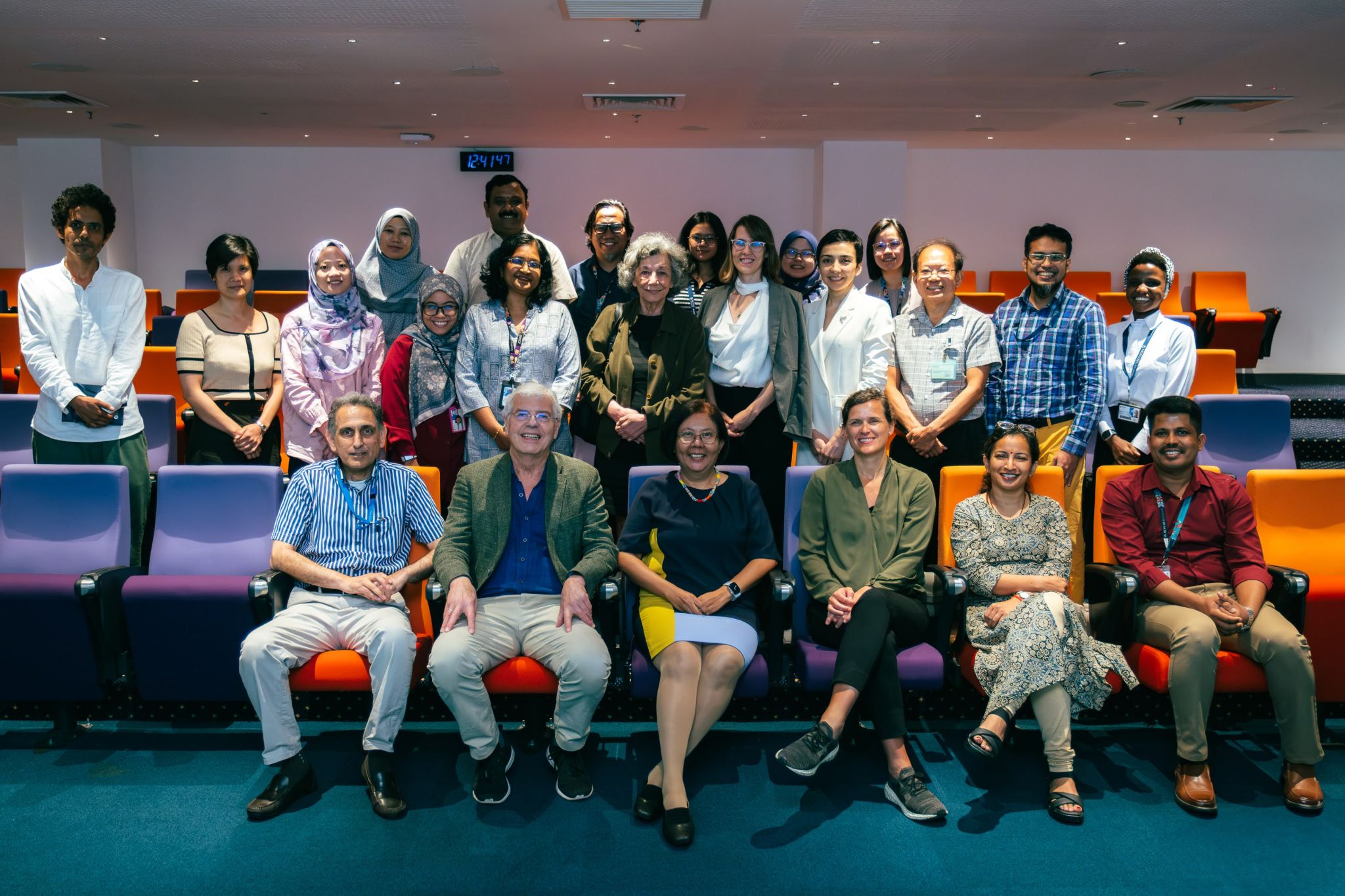 Group photo of diverse international researchers and professionals seated and standing in a brightly lit auditorium