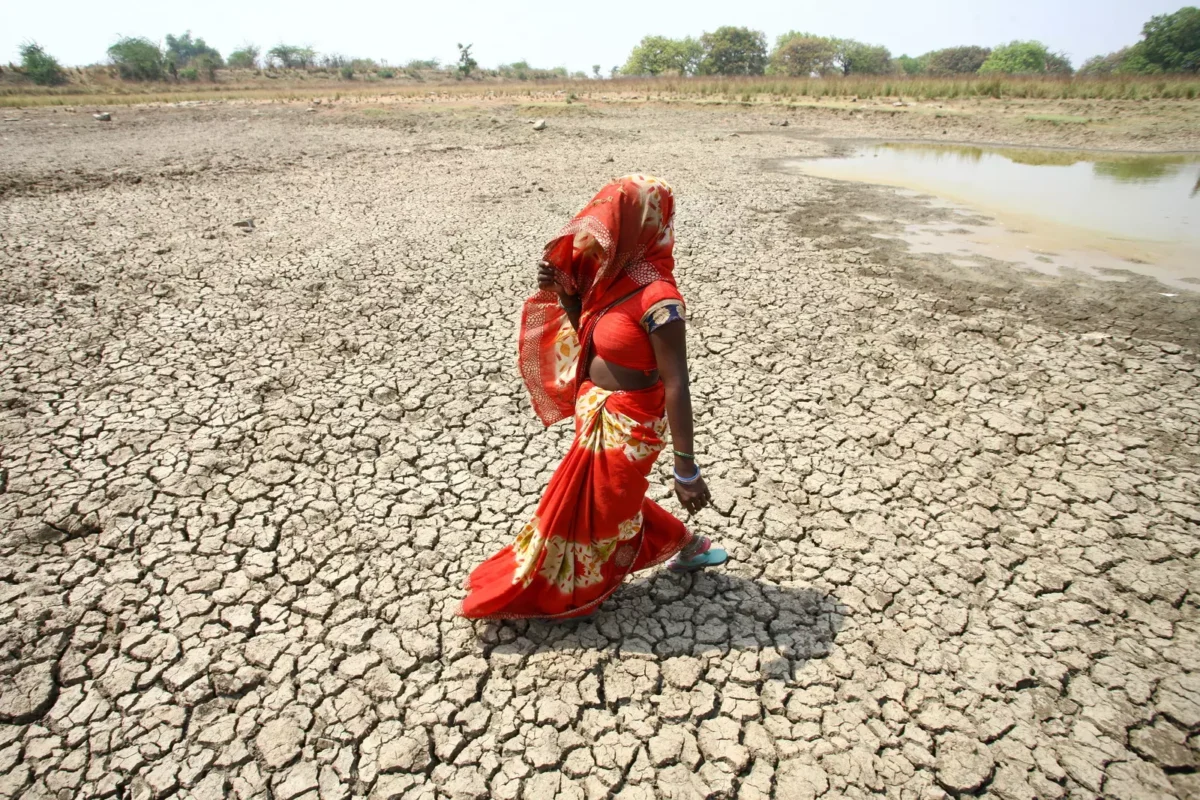 A woman in a red sari walks across a dry, cracked landscape near a shrinking water source, reflecting severe drought conditions.