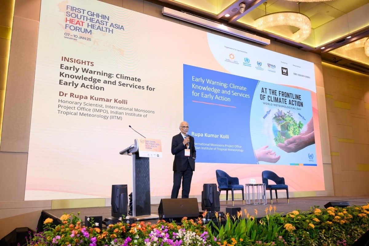 Dr. Rupa Kumar Kolli stands on a stage speaking at a podium during the First GHHIN Southeast Asia Heat Health Forum 2025 in Singapore. Behind him, a large screen displays the title of his talk: “Early Warning: Climate Knowledge and Services for Early Action.” The stage is decorated with flowers,