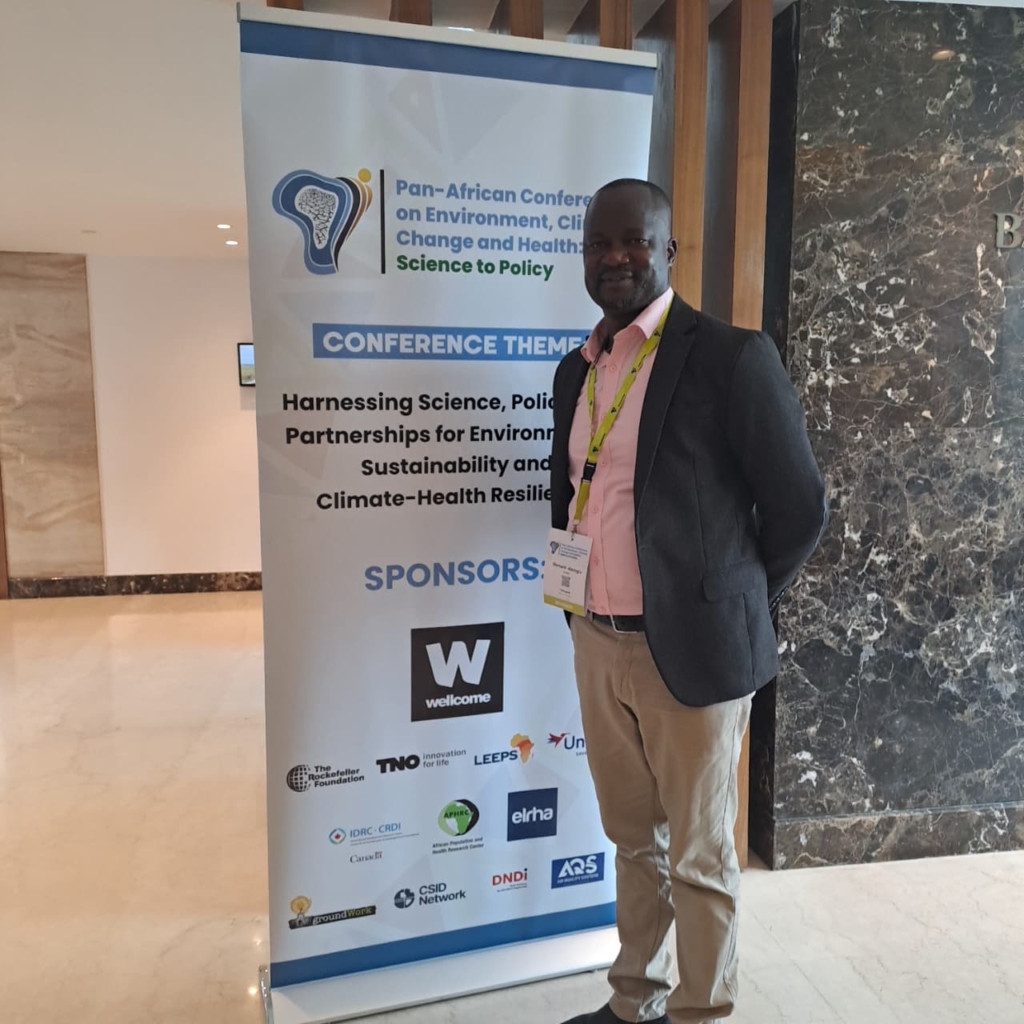 A photo of Dr. Bernard Abong'o, standing in front of a conference banner for the Pan-African Conference on Environment, Climate Change and Healthcare.