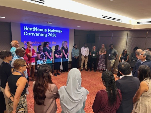 A diverse group of people stands in a semi-circle during a meeting, with a screen displaying