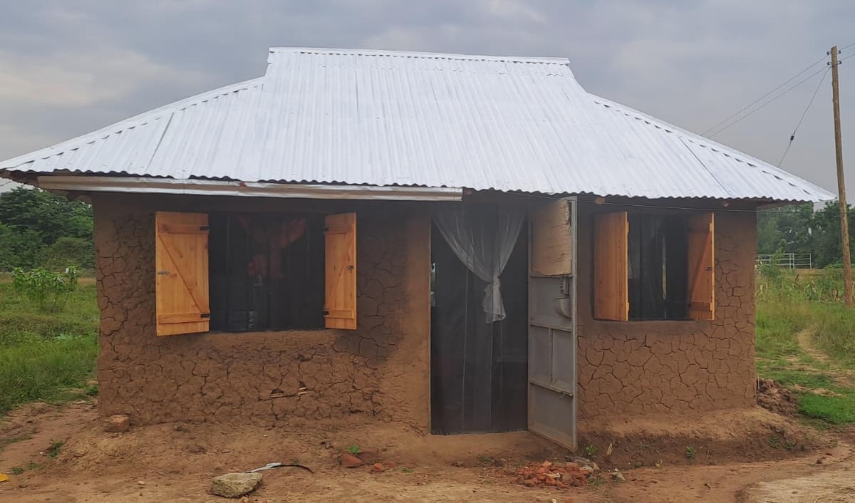 A single house with mud walls, a white-painted tin roof, wooden shutters and net curtains