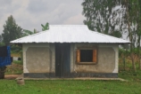 A photograph of a single village house surrounded by grass with a door screen, eave-screen, window curtain and white tin roof