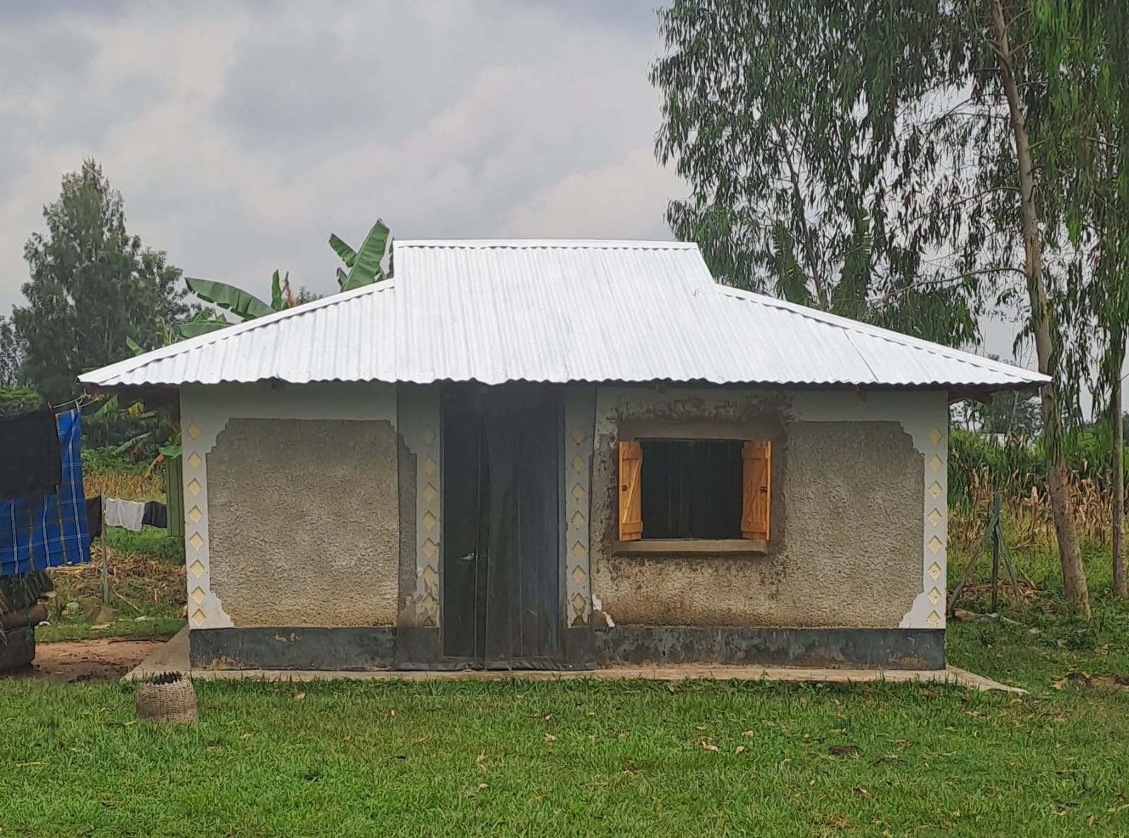 A photograph of a single village house surrounded by grass with a door screen, eave-screen, window curtain and white tin roof