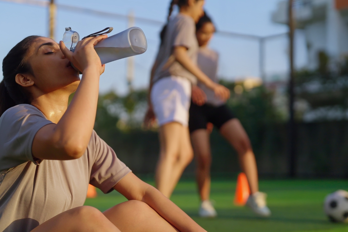 Person sitting on a sports field drinks from a water bottle after training, while other players practice with cones and a football in the background.