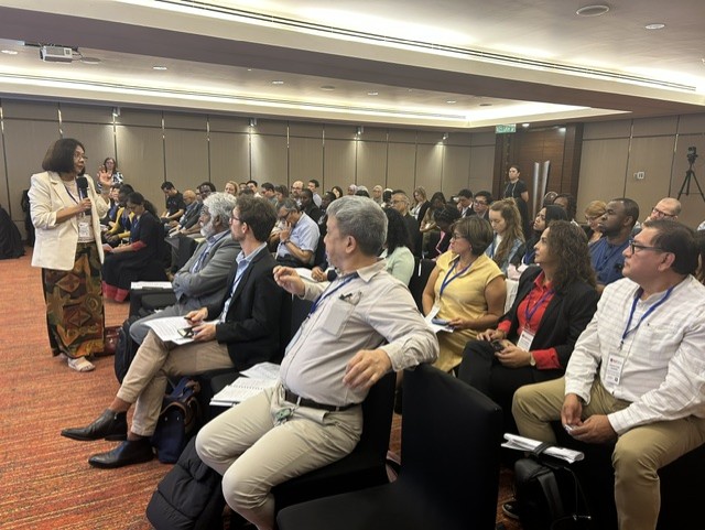 Tin Tin Su stands and speaks to a seated audience in a conference room, with attendees listening and taking notes